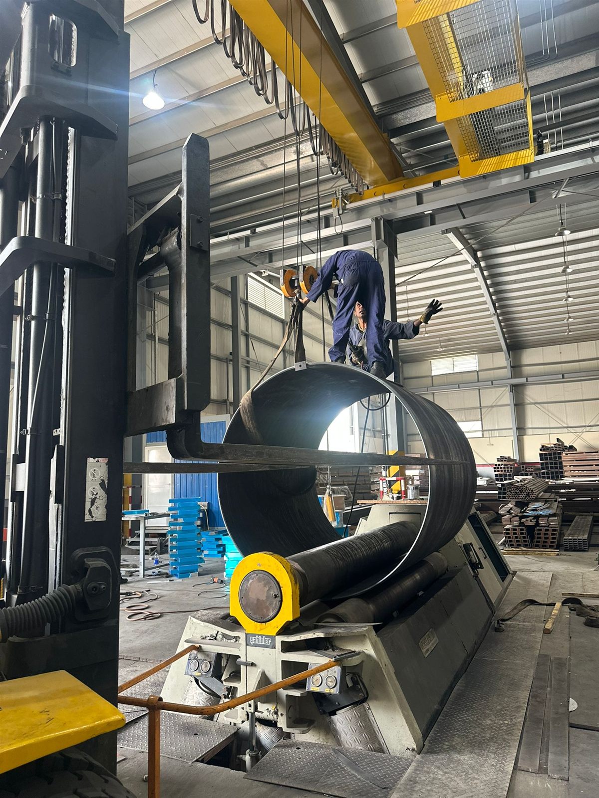 A rolled steel shell lifted off the rolls by crane while a worker guides the sling