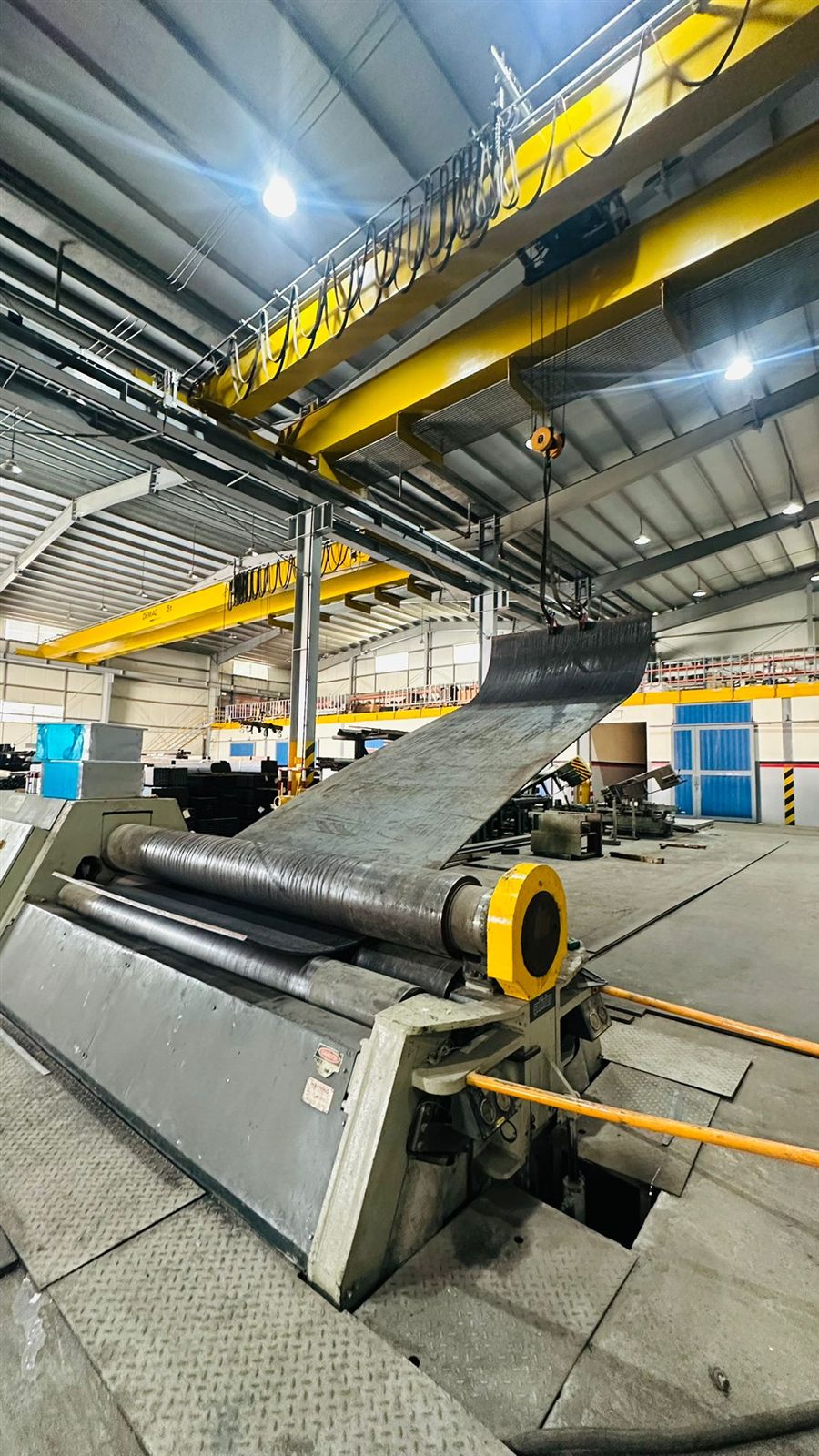 A steel plate being fed into a three-roll plate bending machine under an overhead crane